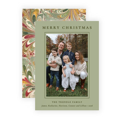 A personalized Christmas family photo card with a sage green border and a central family portrait, shown in front of a matching patterned envelope with red, gold, and green marbled design on a white background.