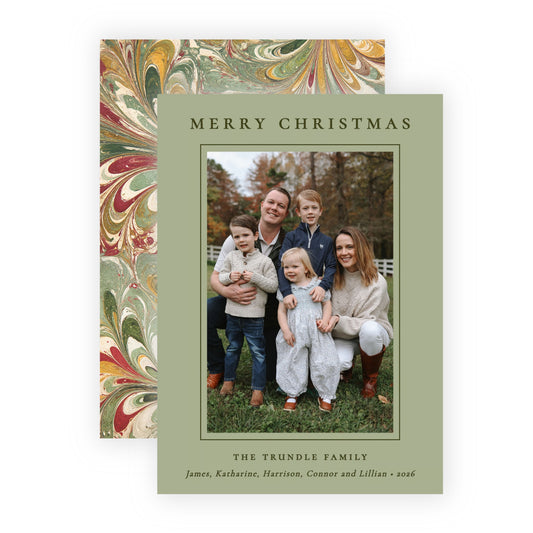 A personalized Christmas family photo card with a sage green border and a central family portrait, shown in front of a matching patterned envelope with red, gold, and green marbled design on a white background.
