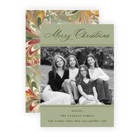 A personalized Christmas family card with a black-and-white photo of four women, set against a sage green background with elegant cursive text reading 'Merry Christmas' and printed family names at the bottom. A matching patterned envelope or backing card with red, gold, and green marbled swirls is visible behind the card on a white background.