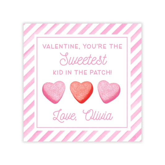 The image is a Valentine's Day card with a pink and white striped border. The card features three heart-shaped candies, each with a different color: pink, red, and pink. The text on the card reads, "Valentine, you're the Sweetest Kid in the Patch! Love, Olivia".